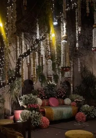 Mehndi stage backdrop with hanging jasmine and pearl strings, fairy lights, and a low cushioned seat framed by floral arrangements