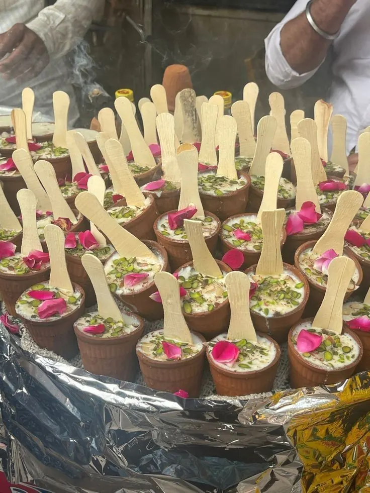 Clay kulhar cups of pistachio-and-rose-petal phirni with wooden spoons at a dessert station